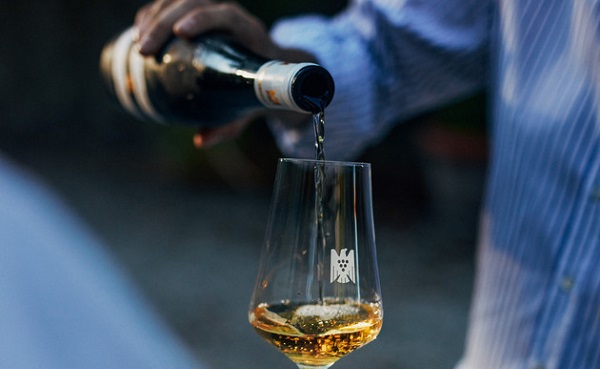 Ein Glas wird mit goldfarbenem Getränk gefüllt, während eine Person eine Flasche mit einem schwarzen Etikett über dem Glas hält, um zu gießen. Der Hintergrund bleibt unscharf.