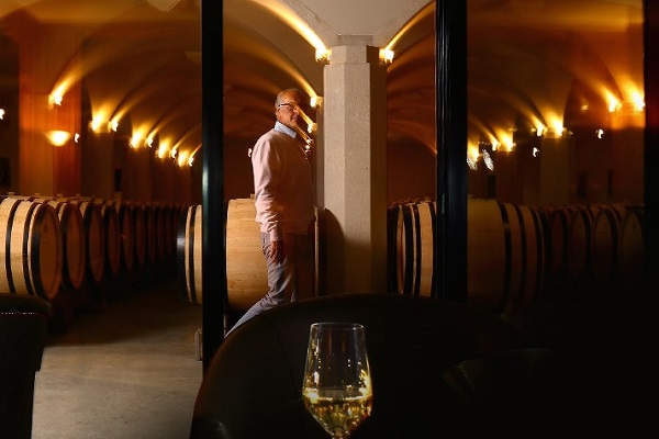 Ein Mann in heller Kleidung steht in einem Weinkeller, umgeben von großen Holzfässern. Ein Glas Wein steht auf einem Tisch im Vordergrund. Die Beleuchtung schafft eine warme Atmosphäre.