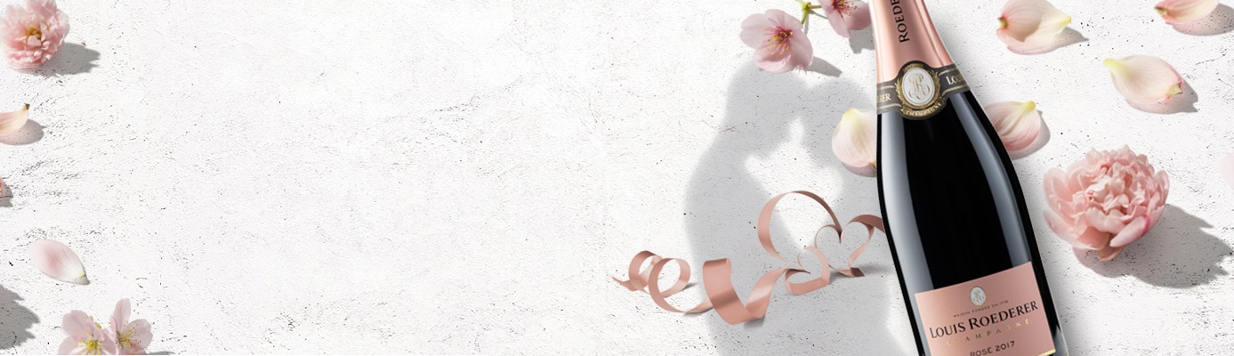 Eine Flasche Louis Roederer Rosé Champagner steht im Vordergrund, umgeben von zarten rosa Blütenblättern und einer spiralförmigen Ripsbandanordnung, auf einer hellen, strukturierten Oberfläche.