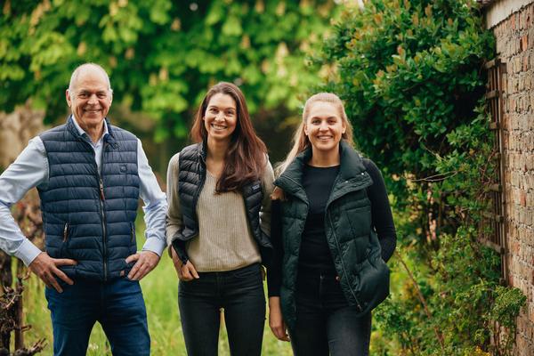 Eine Gruppe von drei Personen steht in einer grünen Umgebung. Sie tragen Westen und lächeln in die Kamera. Im Hintergrund sind Bäume und eine Mauer aus Steinen sichtbar.