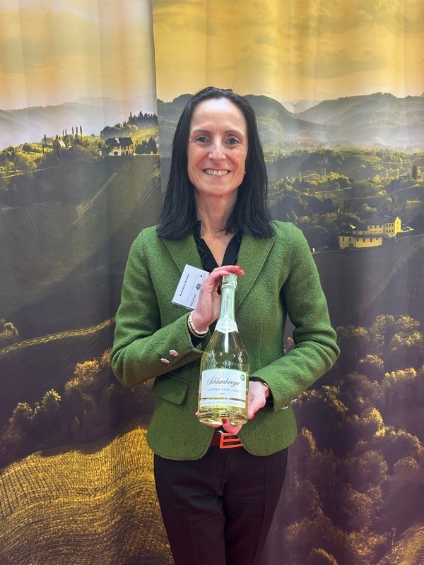 Eine Frau mit langen Haaren trägt einen grünen Blazer und hält eine Flasche in der Hand. Im Hintergrund sind sanfte Hügel und eine malerische Landschaft mit Weinbergen zu sehen.