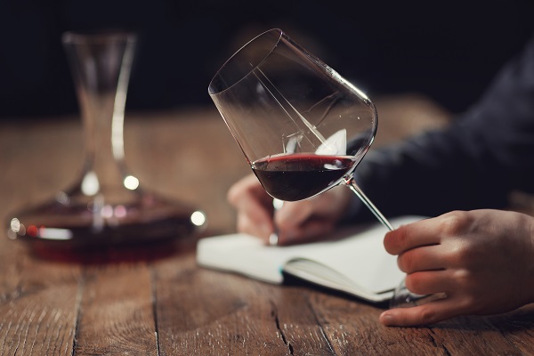 Ein Glas Rotwein wird gehalten, während eine handschriftliche Notiz auf einem Notizblock gemacht wird. Im Hintergrund steht eine Dekantierkaraffe auf einem Holztisch.
