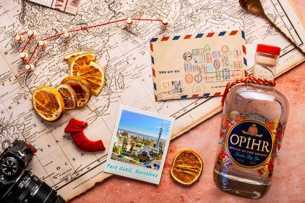 Eine Flasche OPHIR Gin liegt auf einer Landkarte, umgeben von getrockneten Orangenscheiben, einem Postkartenmotiv von Park Güell in Barcelona, einer roten Schnur und einer Briefmarkenmarkierten Postkarte.