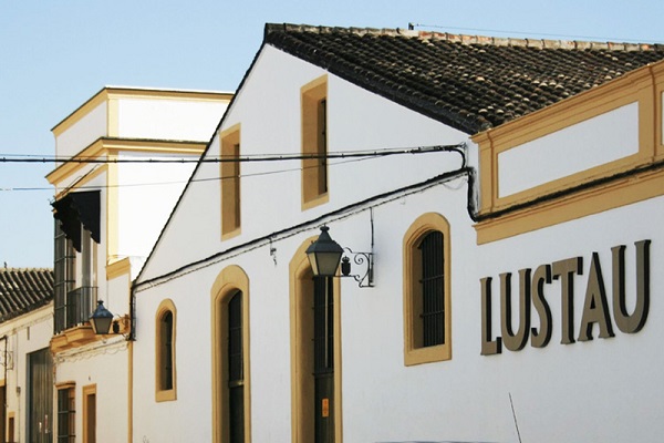Ein historisches Gebäude mit weißer Fassade und gelben Details. Es zeigt den Schriftzug -LUSTAU- und hat eine Laterne sowie Fenster mit farbigen Vorhängen. Der Himmel ist klar.