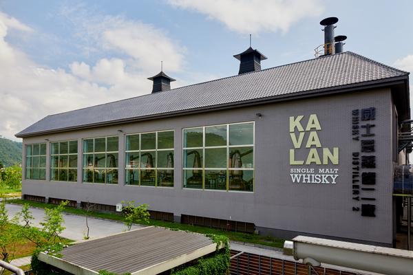 Ein modernes Gebäude mit großen Fenstern, das Kavalan Single Malt Whisky produziert. Im Hintergrund sind sanfte Hügel und ein blauer Himmel sichtbar, während im Vordergrund eine grüne Landschaft zu sehen ist.