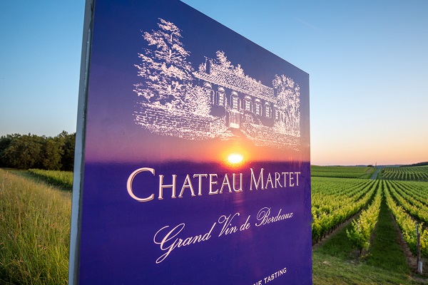 Ein großes Schild mit der Aufschrift -Chateau Martet- steht im Vordergrund, während im Hintergrund Weinreben und ein Sonnenuntergang über den Weinbergen in Bordeaux sichtbar sind.