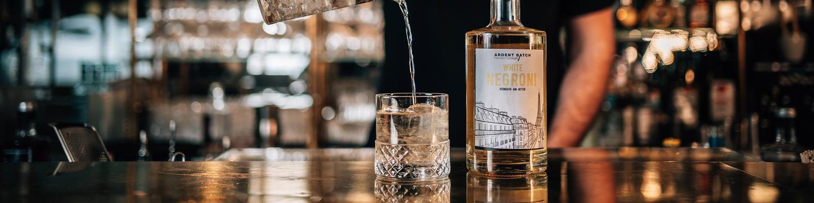 Ardent Batch Ein Glas mit Eis steht auf der Theke, während ein klarer Getränkefluss aus einer Flasche, die -White Negroni- etikettiert ist, in das Glas gegossen wird. Im Hintergrund sind verschwommene Barutensilien sichtbar.