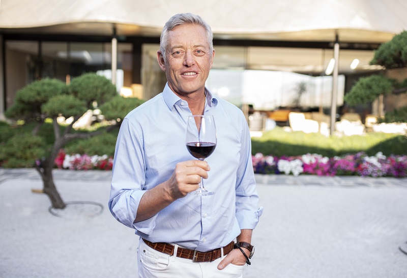 Ein Mann hält ein Glas Rotwein in einer entspannenden Umgebung mit modernen Möbeln und bunten Blumen im Hintergrund. Er trägt ein hellblaues Hemd und lächelt freundlich.