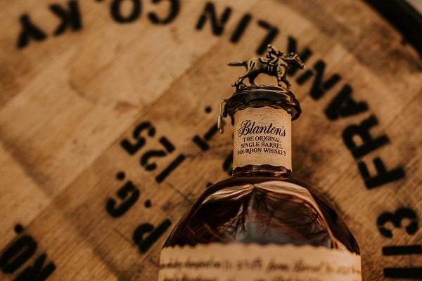 Eine Flasche Blanton-s Bourbon mit einem Pferdchen auf dem Verschluss steht auf einem Holzfass. Der Etikett zeigt den Namen und die Beschreibung des Whiskeys deutlich.