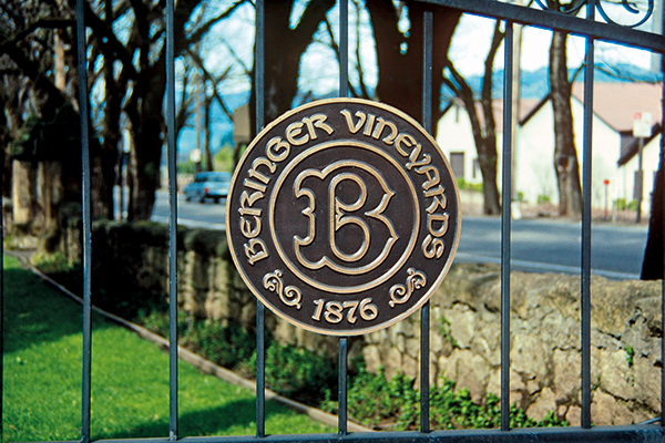 Ein rundes Schild aus Metall mit dem Schriftzug -Beringer Vineyards- und der Jahreszahl -1876- ist an einem metallenen Tor angebracht. Im Hintergrund sind Bäume und Gebäude sichtbar.