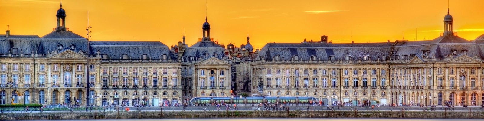 Historische Gebäude mit klassischen Fassaden stehen am Ufer, während eine Straßenbahn vorbeifährt. Ein Sonnenuntergang taucht den Himmel in warme Farben und Menschen flanieren entlang des Wassers.