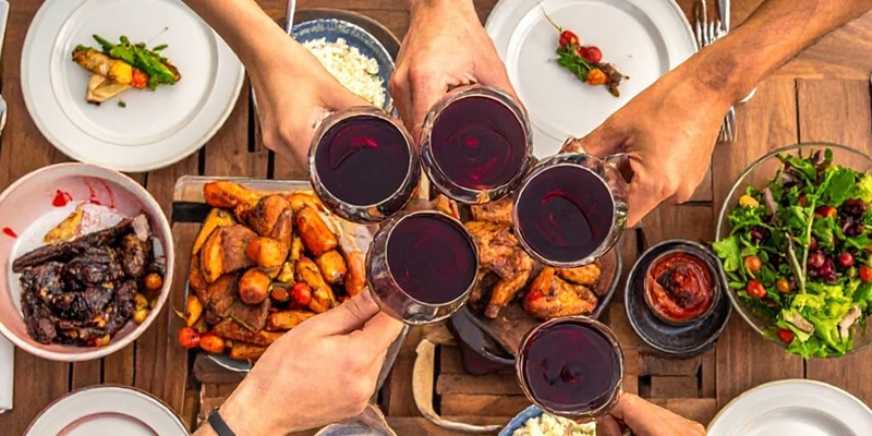 Eine Gruppe von Händen hebt Weingläser und Flaschen an, während sie sich um einen Tisch mit verschiedenen Speisen wie Pasta, Salat, Würstchen und gegrilltem Gemüse versammeln.