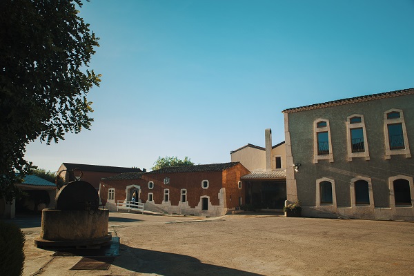 Ein Weingut mit mehreren Gebäuden in verschiedenen Farben, darunter rot und grau. Im Vordergrund befindet sich eine große Steinstruktur. Der Himmel ist blau und klar.