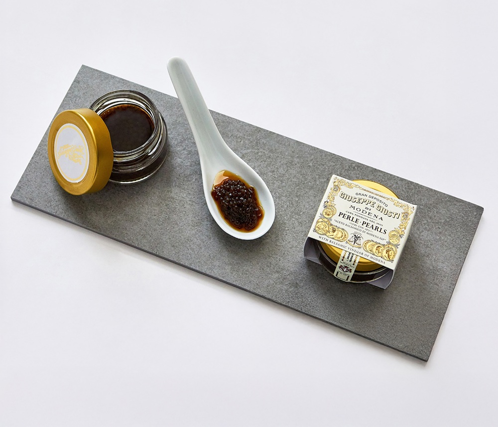 Ein rechteckiger, grauer Steinplatte zeigt ein Glas mit goldenem Deckel, einen Löffel mit dunklen Perlen und eine dekorative Schachtel, die Perlen mit Balsamico-Essig enthält.