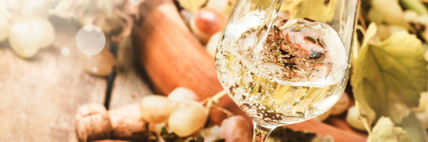 Ein Glas mit sprudelndem, hellem Wein steht im Vordergrund. Im Hintergrund liegen Weintrauben und Weinblätter auf einem Holztisch, was eine entspannte Atmosphäre schafft.