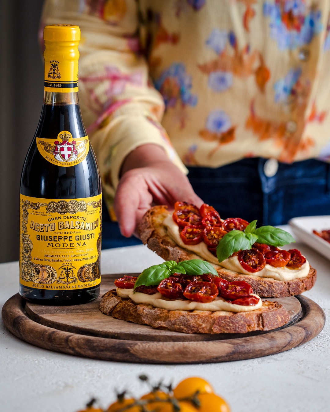 Eine Flasche Balsamico-Essig steht neben zwei belegten Brotscheiben mit sonnengetrockneten Tomaten und Basilikum auf einem Holzbrett. Eine handvoll Kirschtomaten ist im Vordergrund sichtbar.