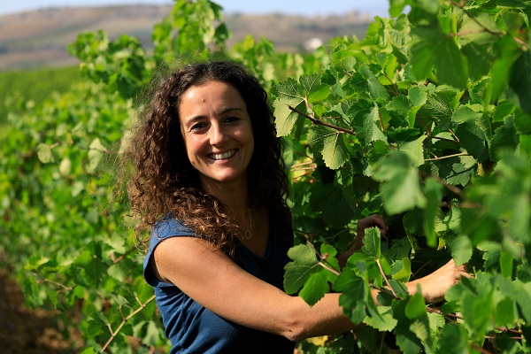 Eine Frau mit lockigem Haar lächelt, während sie in einem Weinberg steht und mit ihren Händen an den Weinblättern arbeitet. Im Hintergrund sind sanfte Hügel zu sehen.