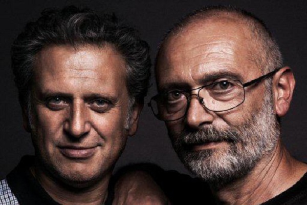 Zwei Männer stehen eng beieinander und lächeln in die Kamera. Der eine hat lockiges, dunkles Haar und der andere eine Glatze mit Brille und grauem Bart. Der Hintergrund ist dunkel.