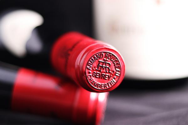 Eine Rotweinflasche mit einer roten Kapsel im Vordergrund. Die Kapsel hat einen geprägten Schriftzug. Im Hintergrund ist eine weitere Flasche unscharf sichtbar, alles vor einem dunklen Untergrund.