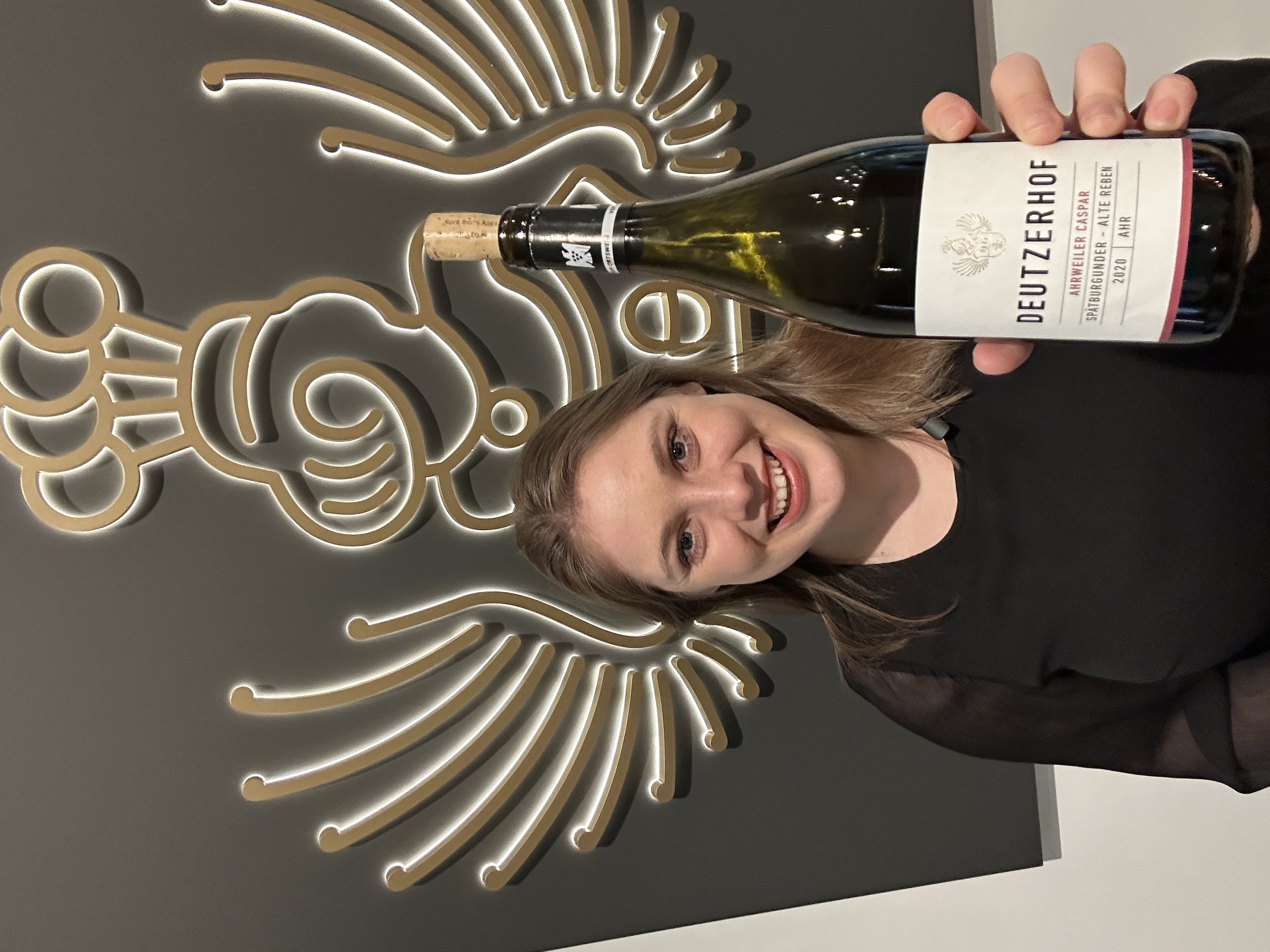 Eine Person hält eine Flasche Rotwein mit dem Etikett -Deutzerhof Ahrweiler Caspar Spätburgunder - Alte Reben 2020- vor einer Wand mit einer stilisierten Koch- und Flügelgrafik.