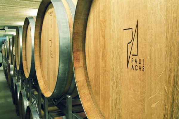 Holzfässer stehen auf Regalen in einem Weinkeller. Die Fässer sind mit einem Logo und dem Namen -PAUL ACHS- versehen. Der Raum hat eine gedämpfte Beleuchtung.