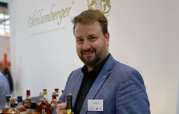 Ein Mann mit kurzer, brauner Haar styl und Bart steht lächelnd, während er eine Flasche in der Hand hält. Im Hintergrund sind mehrere Flaschen auf einem Tisch zu sehen.