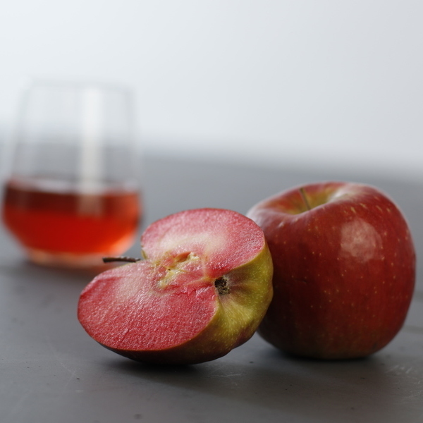 Ein roter Apfel liegt auf einem grauen Untergrund, mit einem halbierten Apfel daneben. Im Hintergrund steht ein Glas mit einer roten Flüssigkeit.