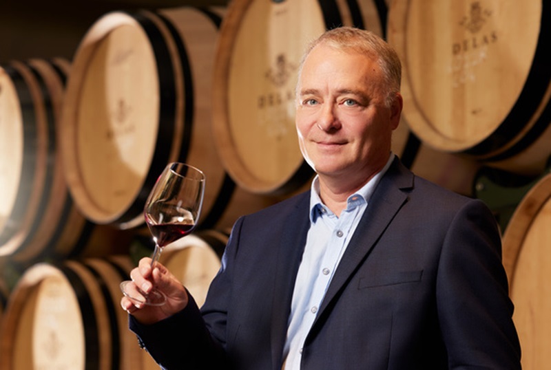 Ein Mann in einem Anzug hält ein Weinglas mit rotem Wein in der Hand. Im Hintergrund sind Holzfässer zu sehen, die angeordnet sind.