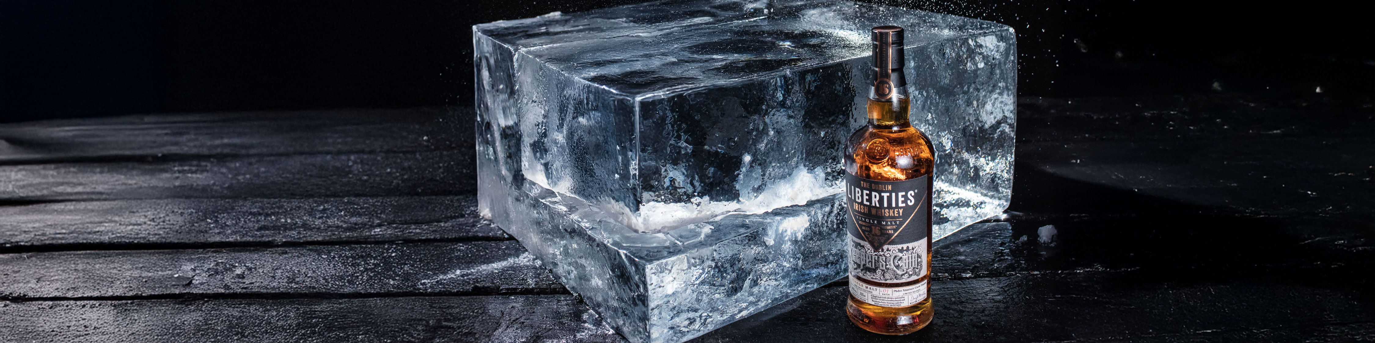 Ein großes, klarer Eisblock steht auf einer dunklen Oberfläche. Daneben befindet sich eine Flasche Irish Whiskey mit einer ausgefallenen Etikettgestaltung, umgeben von leichtem Sprühnebel.