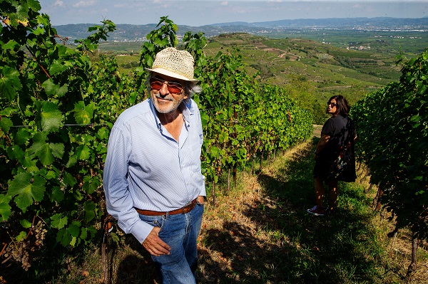 Ein Mann mit Sonnenhut steht in einem Weinberg, lächelnd und entspannt. Im Hintergrund ist eine Frau zu sehen, die im Weinberg steht, während die Landschaft bergig und grün ist.