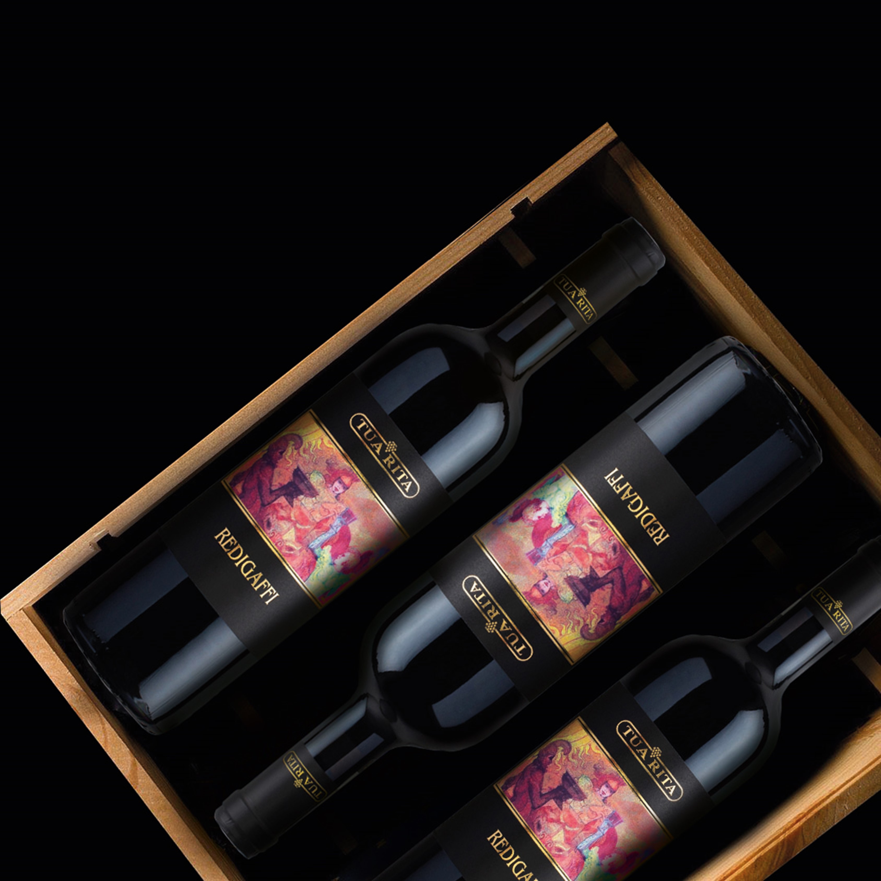 Original Holzkiste Redigaffi Vier Weinflaschen liegen in einer eleganten Holzkiste. Die Flaschen haben schwarze Etiketten mit bunten, kunstvollen Designs. Die Umgebung ist dunkel gehalten, was den Fokus auf die Flaschen lenkt.