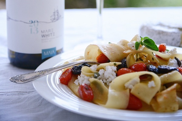 Ein Teller mit frischer Pasta, garniert mit Kirschtomaten, Oliven und Käse, steht neben einer Flasche Weißwein. Der Hintergrund ist hell und natürlich, ideal für ein entspanntes Essen im Freien.