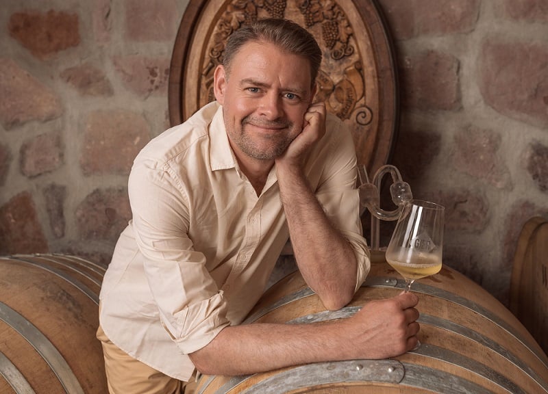 Eine Person lehnt sich entspannt auf ein Holzfass, hält ein Weinglas in der Hand und lächelt. Im Hintergrund sind Weinlagerfässer und eine steinerne Wand sichtbar.