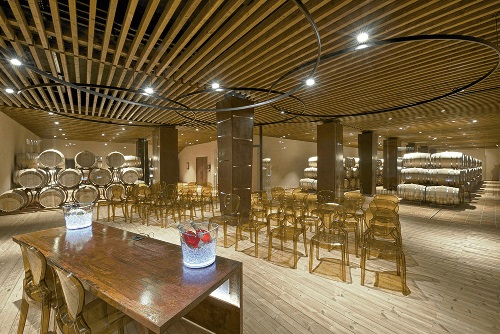 Ein modern gestalteter Weinkeller mit Holzdecke und großen Fässern im Hintergrund. In der Mitte stehen Stühle und ein Tisch mit Gläsern. Die Beleuchtung ist warm und einladend.