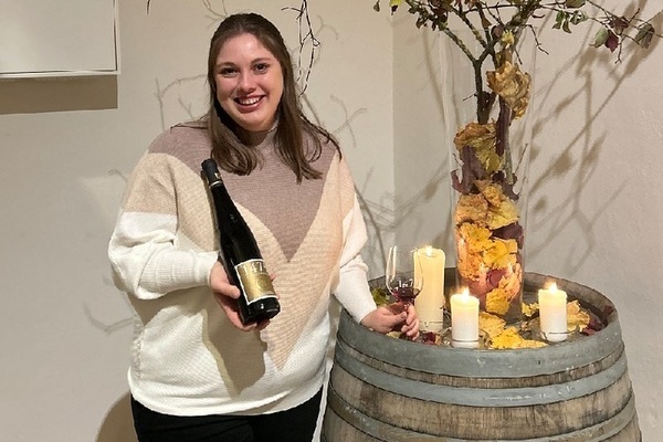 Eine Person hält eine Weinflasche in der Hand und steht lächelnd neben einem Holzfass. Auf dem Fass sind Kerzen und herbstliche Dekorationen platziert.