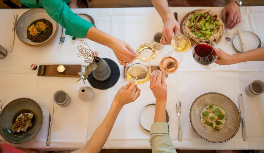Vier Personen stoßen mit Weingläsern an. Der Tisch ist elegant gedeckt mit verschiedenen Gerichten, darunter Salat, Pizza und eine Reis-Schalen. Eine Kerze sorgt für Stimmung.