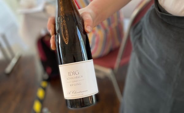 Eine Person hält eine Weinflasche mit einem weißen Etikett. Die Flasche ist ein Riesling mit der Aufschrift -IDIG Longsbach-. Im Hintergrund sind verschwommene Stühle und eine bunte Tasche sichtbar.