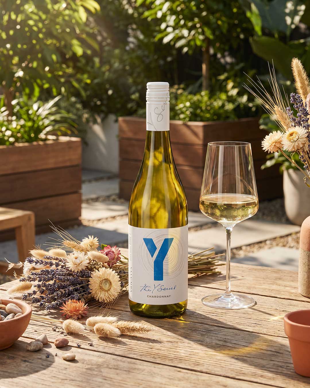 Eine Flasche Chardonnay mit einem blauen Etikett steht neben einem Glas Wein auf einem Holztisch. Im Hintergrund befinden sich Pflanzen und dekorative Trockenblumen zwischen kleinen Steinen.