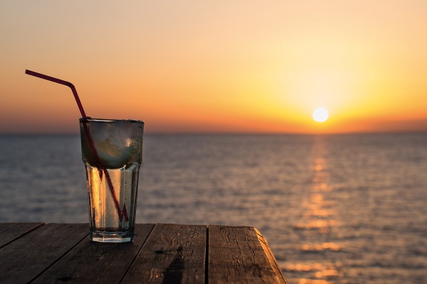 Meer_Strandbar_600 Ein Glas mit einem Getränk und einem Strohhalm steht auf einem Tisch aus Holz. Im Hintergrund leuchtet die Sonne über dem Wasser während der Sonnenuntergangszeit.
