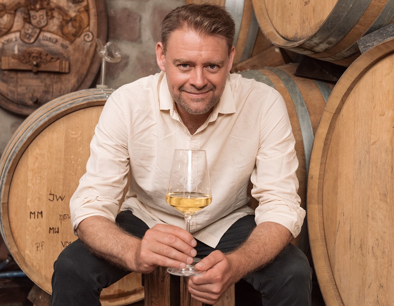 Ein Mann sitzt auf einem Holzfass und hält ein Glas mit einer hellen Flüssigkeit. Im Hintergrund sind weitere Holzfässer sichtbar, was auf eine Weinkellerei hindeutet.