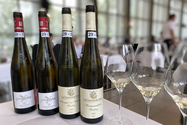 Vier Weinflaschen stehen auf einem Tisch in einem Restaurant, neben zwei Gläsern mit Weißwein. Die Umgebung wirkt elegant und modern mit großen Fenstern im Hintergrund.