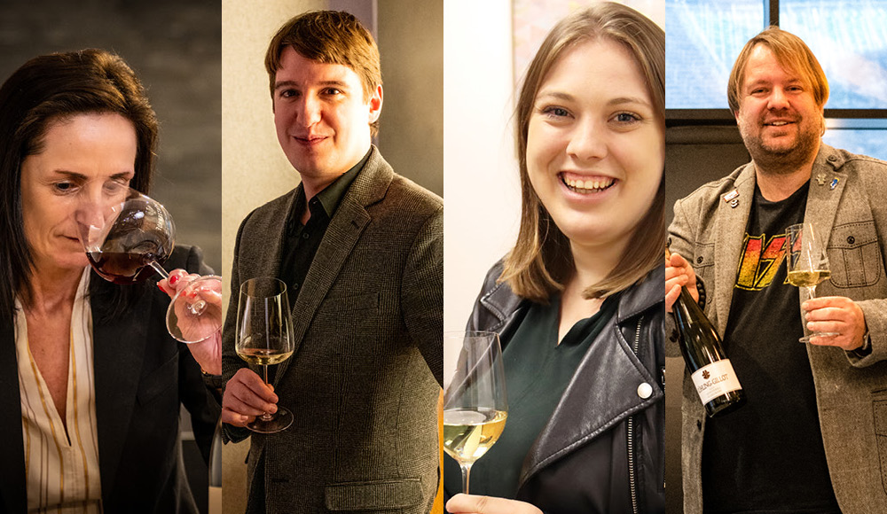 Vier Personen stehen nebeneinander, jede mit einem Weinglas in der Hand. Eine Person riecht an ihrem Glas, während die anderen lächeln und ebenfalls ihre Gläser präsentieren. Der Hintergrund ist gedämpft.