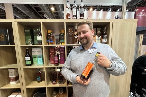 Eine Person hält eine Flasche Spirituosen in der Hand, lächelt und steht vor einem Regal mit verschiedenen Whisky-Flaschen und Verpackungen auf einer Messe.