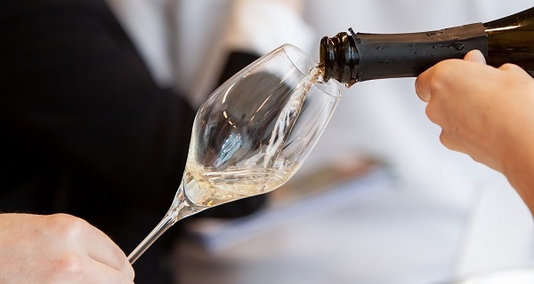 Ein Glas wird mit Schaumwein gefüllt. Eine Hand hält die Flasche, während eine andere Hand das Glas in Position bringt. Der Hintergrund ist unscharf und zeigt eine Umgebung, in der Feierlichkeiten stattfinden.