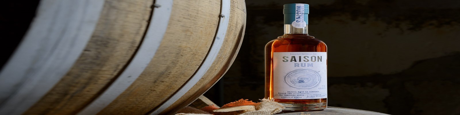 Eine Flasche Saison Rum steht auf einem Holzfass. Im Hintergrund sind weitere Baumstämme und eine rustikale Umgebung zu sehen. Der Fokus liegt auf der Flasche und dem Etikett.