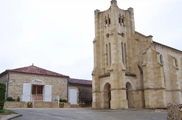 Eine steinerne Kirche mit einem hohen Turm steht im Vordergrund, während ein niedrigerer, angrenzender Bau mit einem Satteldach und Fenstern im Hintergrund sichtbar ist. Die Umgebung ist ländlich und ruhig.