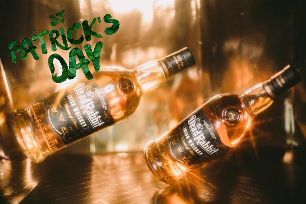 Zwei Flaschen Whiskey stehen schräg auf einer reflektierenden Oberfläche und leuchten im Schein von warmem Licht. Im Hintergrund prangt der Schriftzug -St. Patrick-s Day- in grüner Schrift.