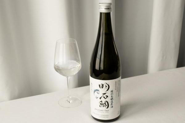 Eine Flasche Sake steht auf einem Tisch neben einem Weinglas, das mit klarem Sake gefüllt ist. Der Hintergrund ist hell, mit Vorhängen aus leichtem Stoff.