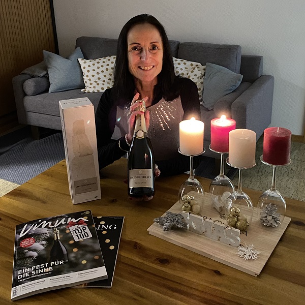 Eine Frau sitzt an einem Tisch, hält eine Flasche Sekt und lächelt. Auf dem Tisch stehen Kerzen, Zeitschriften und eine dekorative Holzplatte mit Weihnachtsmotiven. Im Hintergrund ist ein Sofa sichtbar.