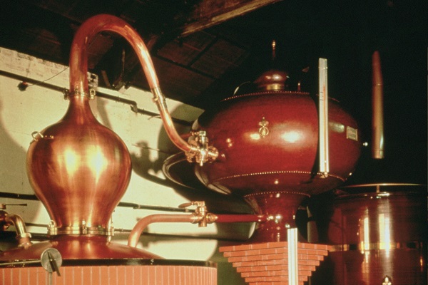 Kupferne Destillationsapparate stehen in einer alten Brennerei. Der eine hat eine geschwungene Form und ein Rohr, das zu einem anderen, runden Behälter führt.